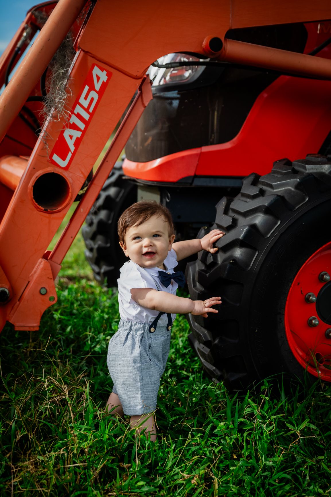 A cheerful baby boy in overalls laughs while touching the tire of a red LA1154 tractor outdoors.