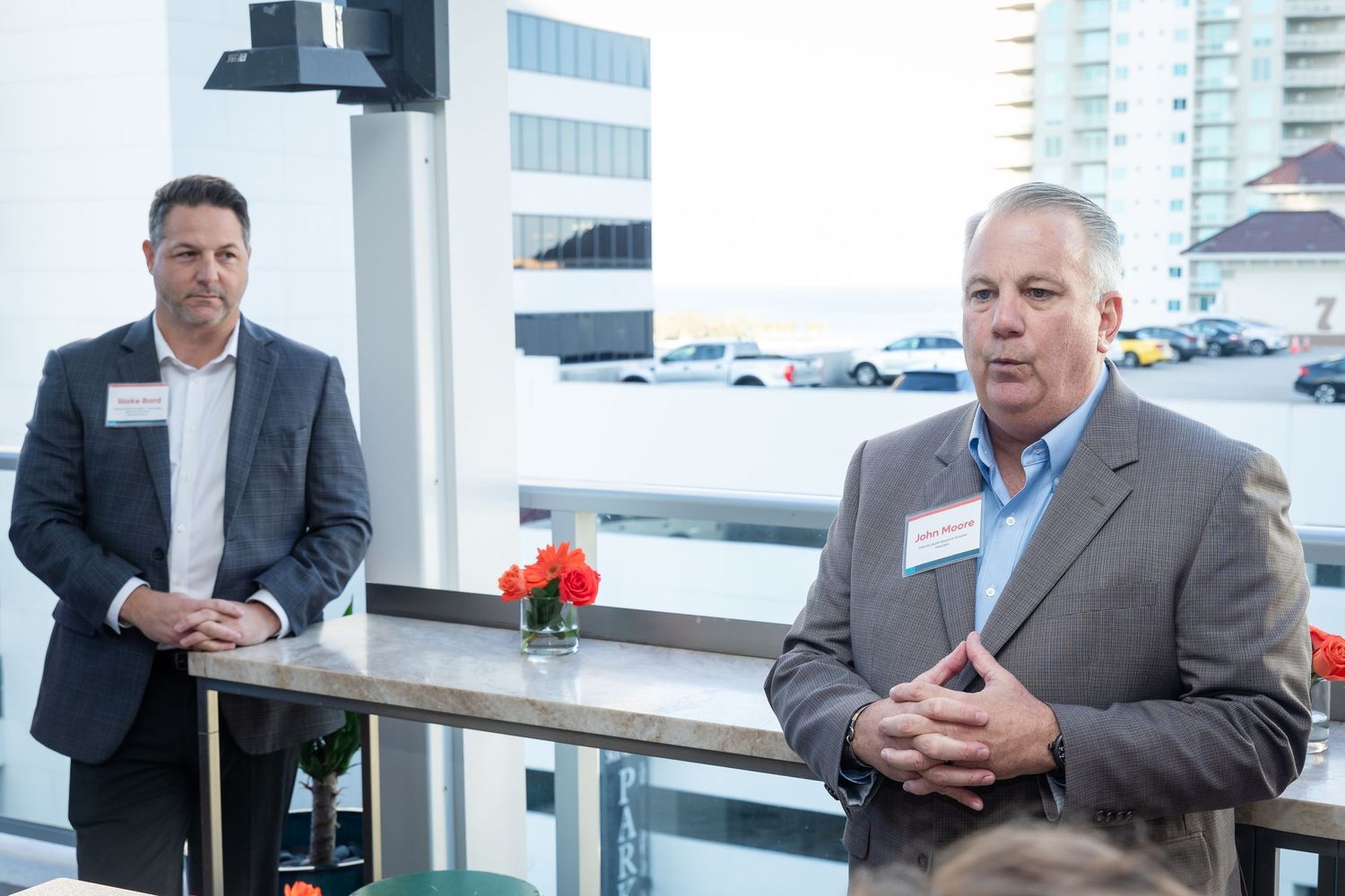 Two businessmen wearing name tags speak to an audience at an outdoor rooftop networking event.