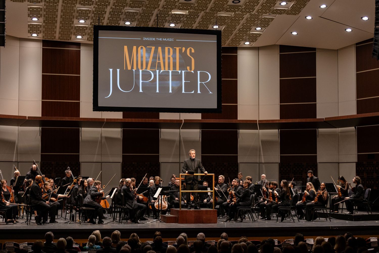 Symphony orchestra performing Mozart's Jupiter beneath a large projected title screen in a concert hall.