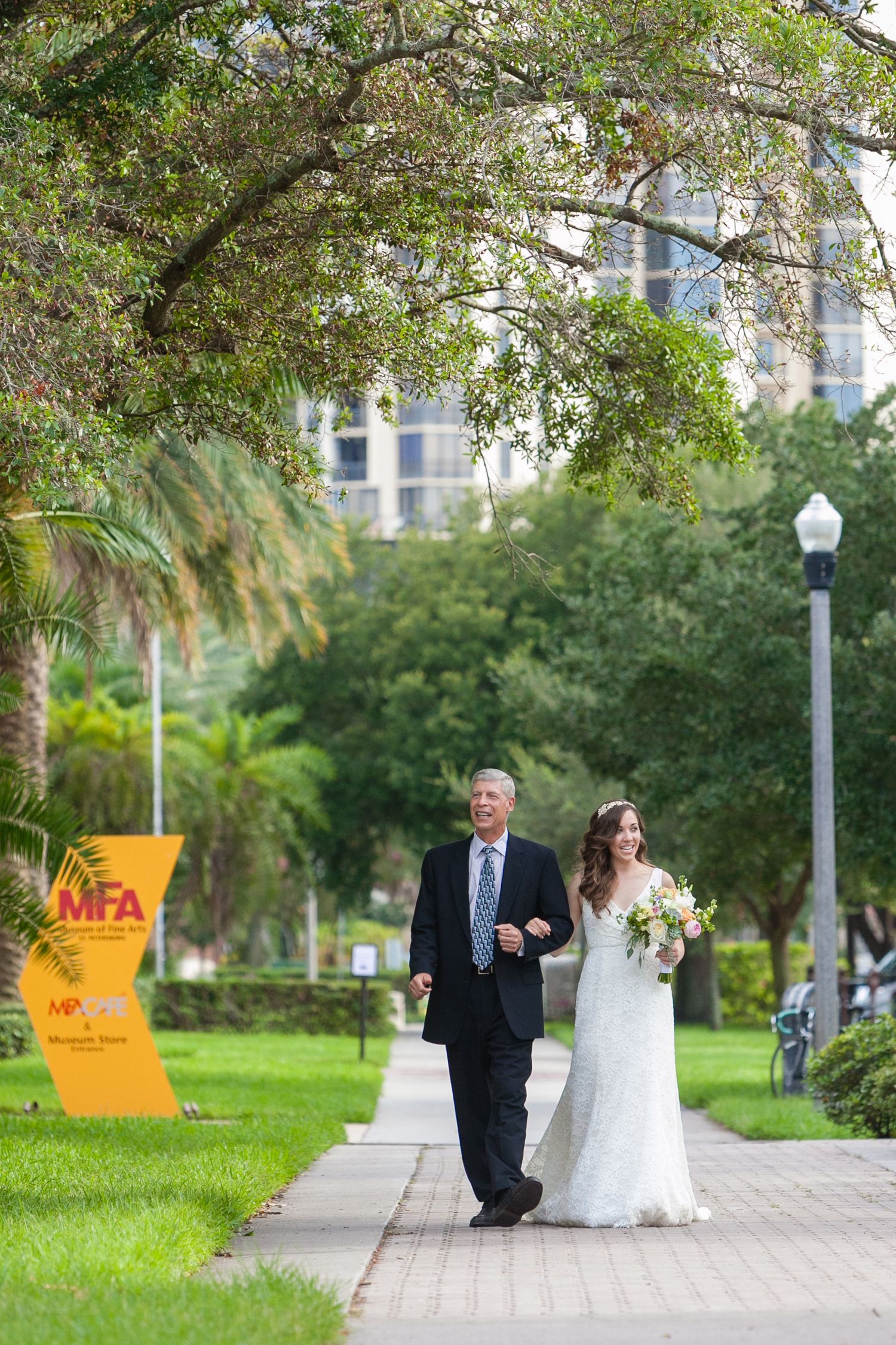 Bride in lace gown walks with father along park pathway near MFA sign on wedding day.