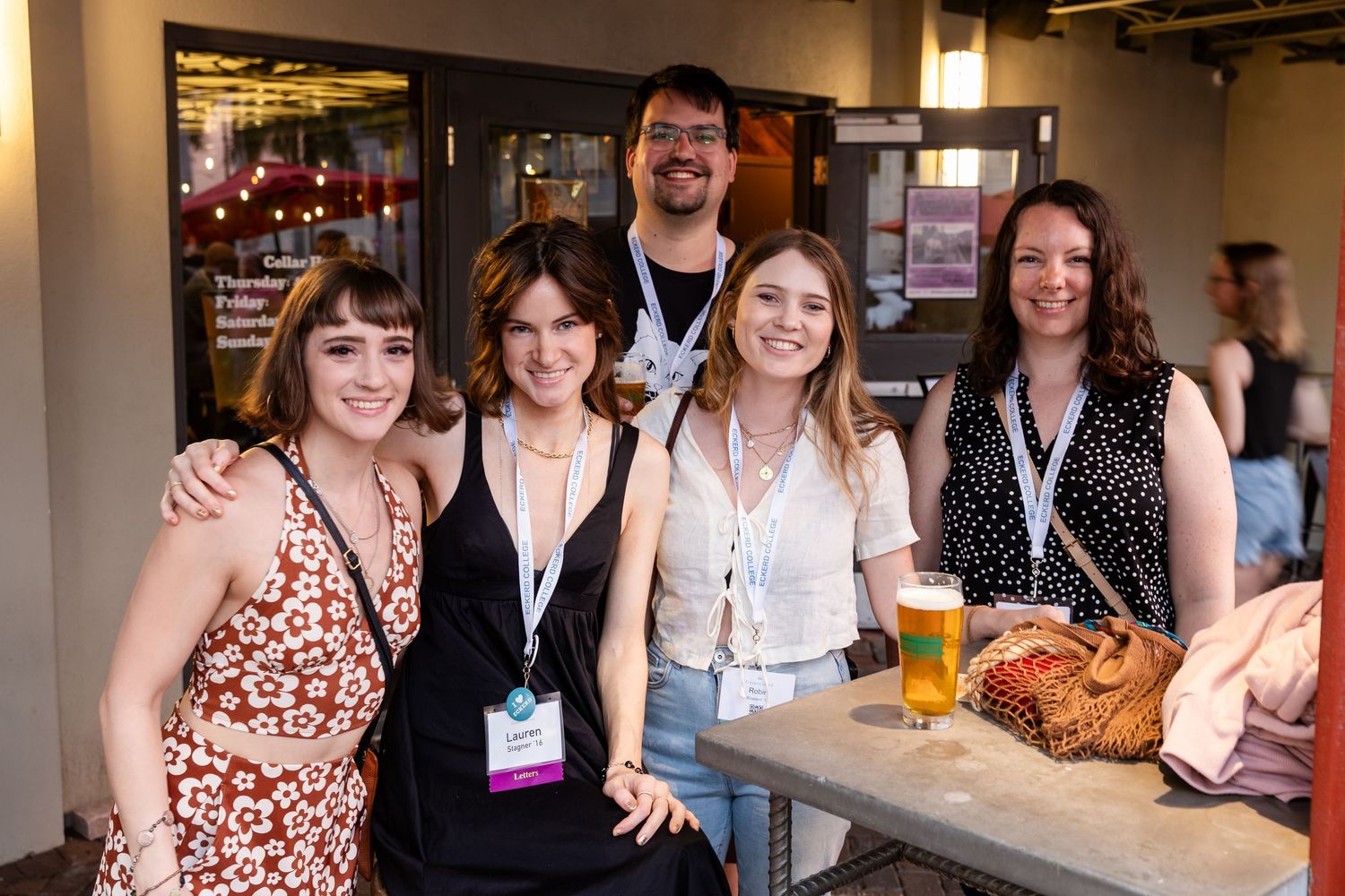 Five people smiling together at an indoor event, with conference lanyards and a glass of beer on a nearby table.