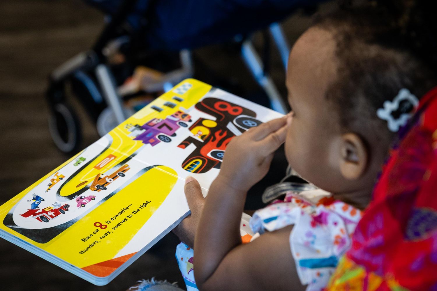 Young toddler girl reading a colorful picture book, pointing at illustrations while seated in a stroller.
