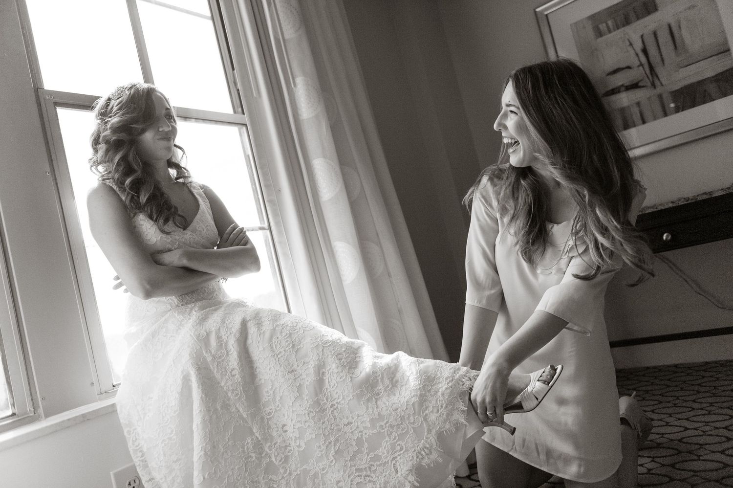 Black and white photo of a bridesmaid helping the bride with her shoes while both share a joyful laugh.