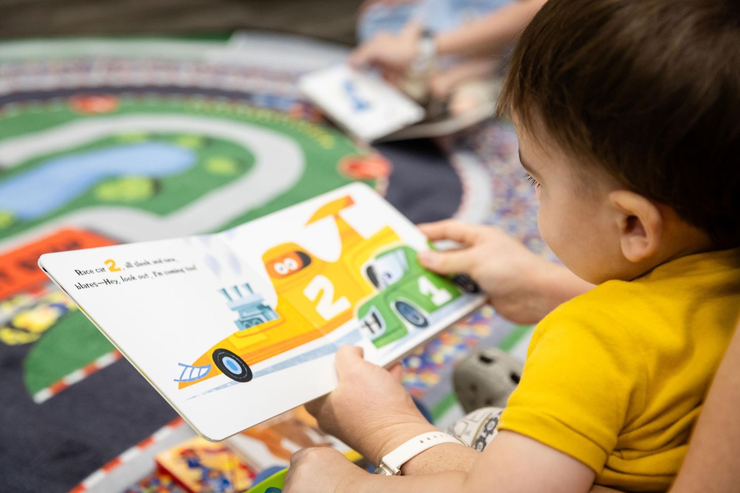 Toddler in yellow shirt reading a colorful children's picture book with cars on a playmat.