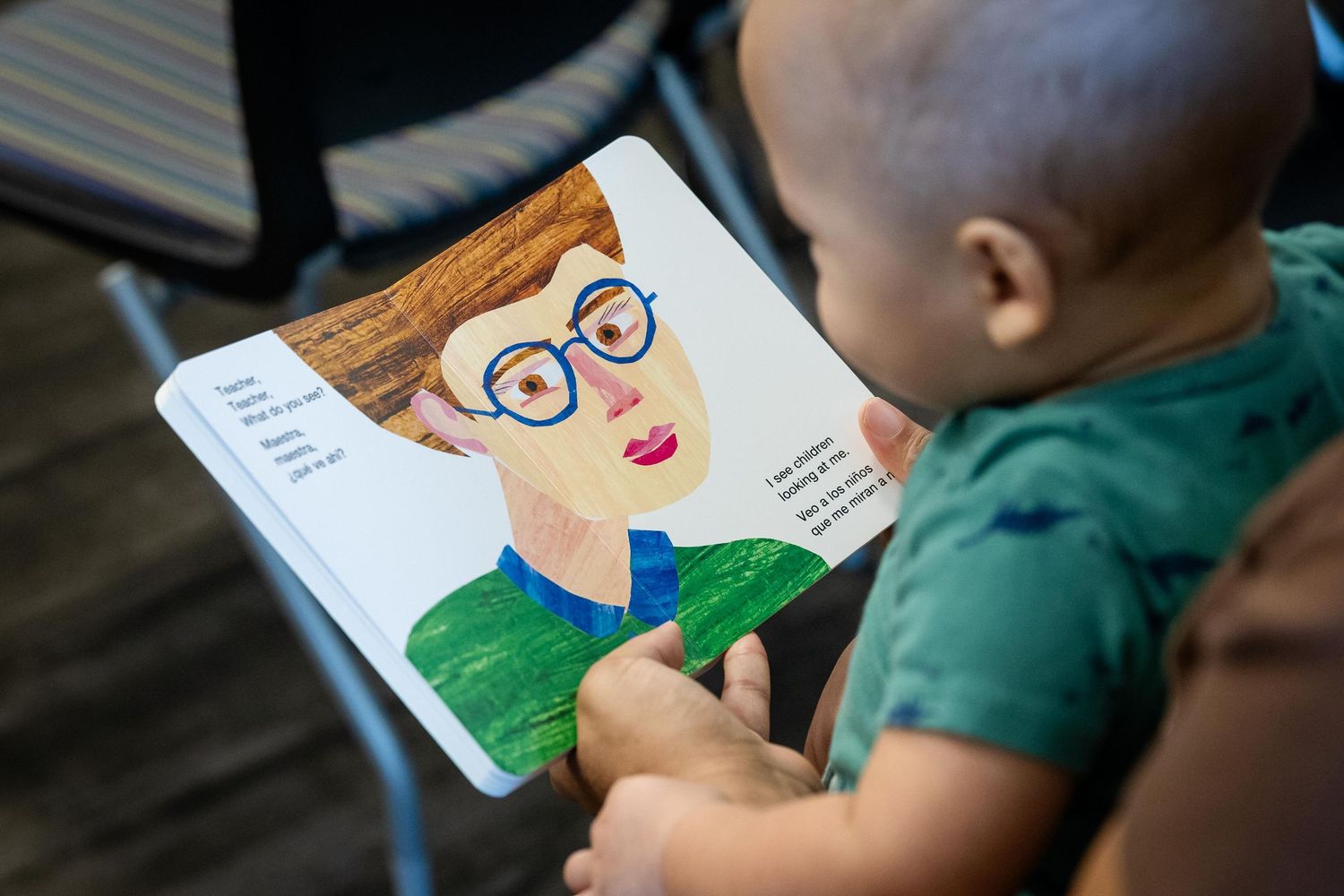 A baby holds a colorful children's board book featuring an illustrated person wearing glasses.