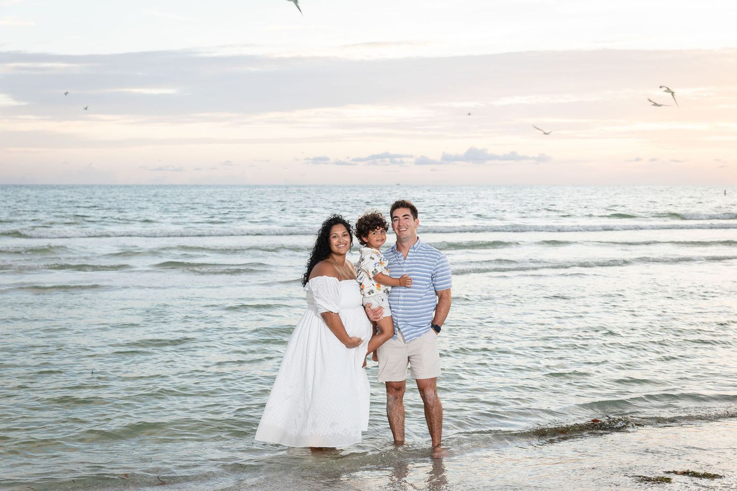 Family of three posing at the beach shoreline at sunset, mother in white dress, father in blue shirt holding toddler.