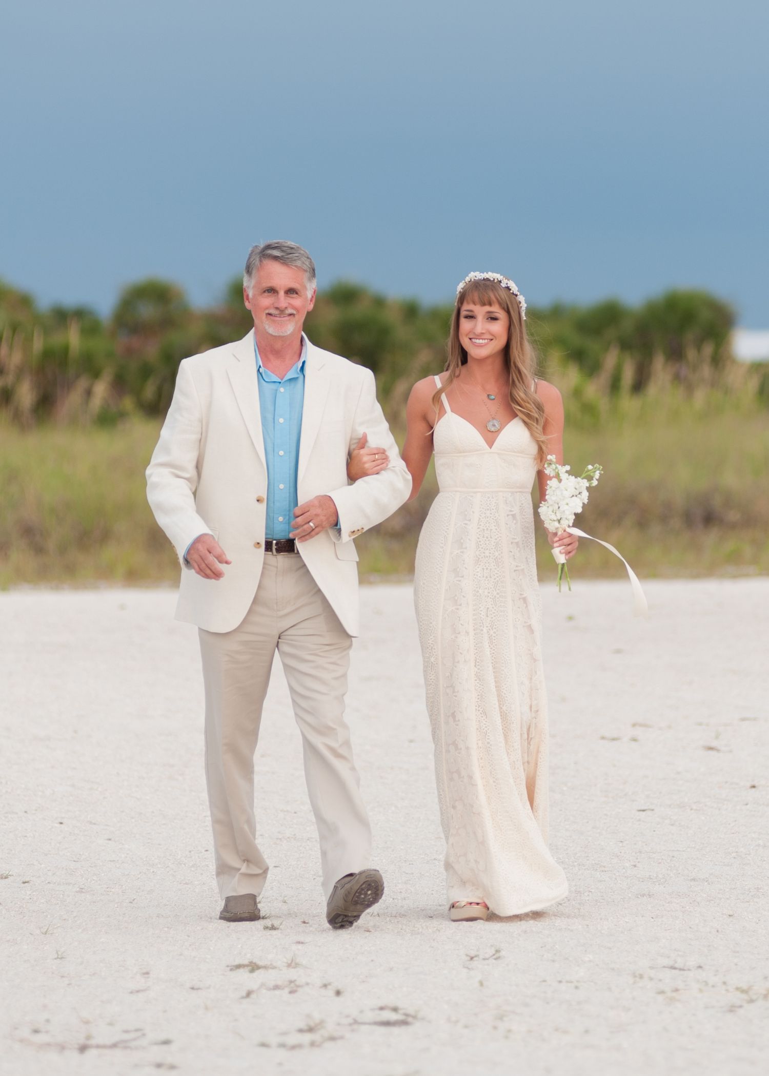 Bride in lace gown holding bouquet walks with man in cream suit on sandy beach during outdoor wedding.