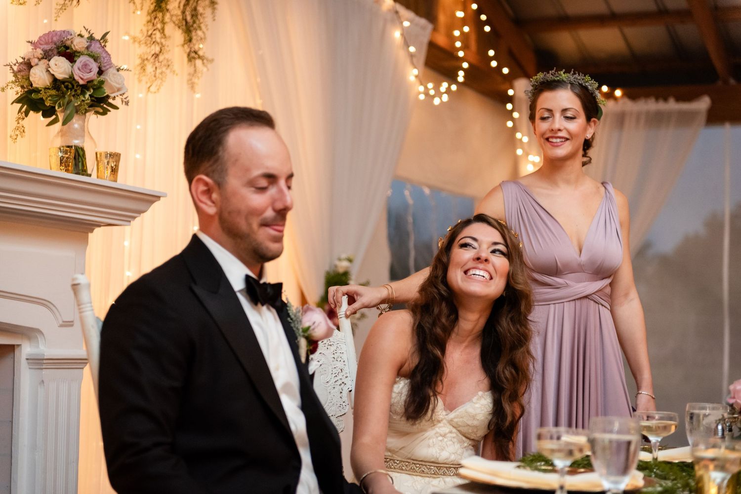 Bridesmaid in lavender dress smiling behind bride and groom at wedding reception dinner table.