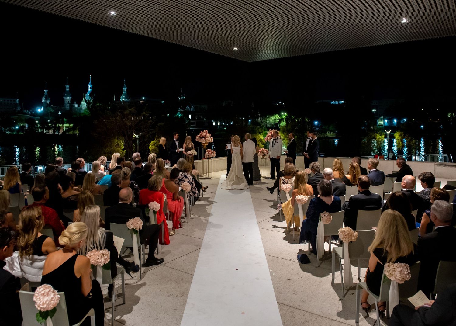Guests seated at an outdoor evening wedding ceremony with city lights in the background.