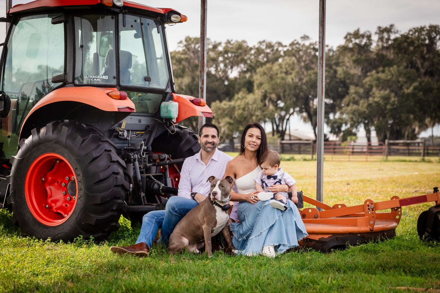 Smiling couple with baby and brown dog sitting beside a red tractor on a lush green farm.