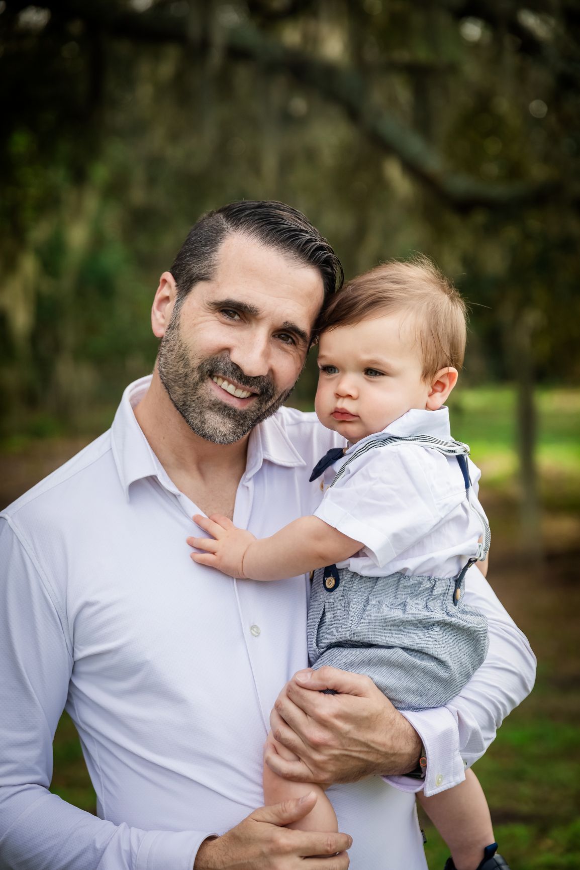 Smiling bearded father holding baby boy in bow tie outfit outdoors.