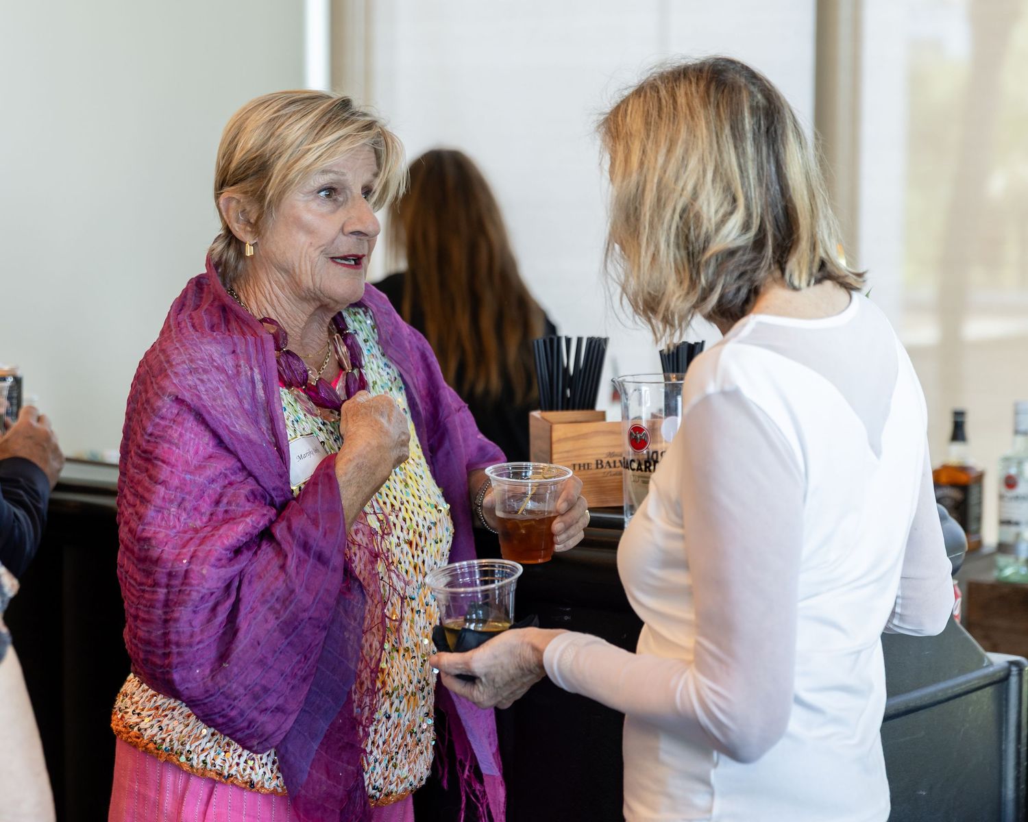 Two women conversing at a social event, one wearing a colorful pink scarf holding a drink.