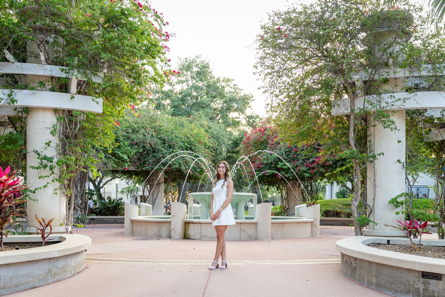 Young woman in white dress poses by elegant fountain courtyard surrounded by lush greenery and colorful flowers.
