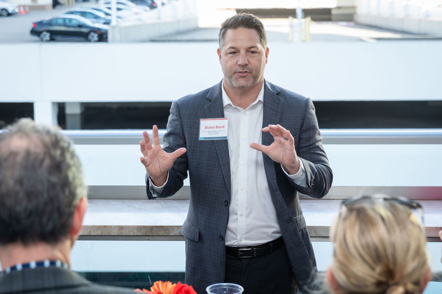 A man in a gray blazer with a name tag speaks gesturing with both hands at a professional business event.
