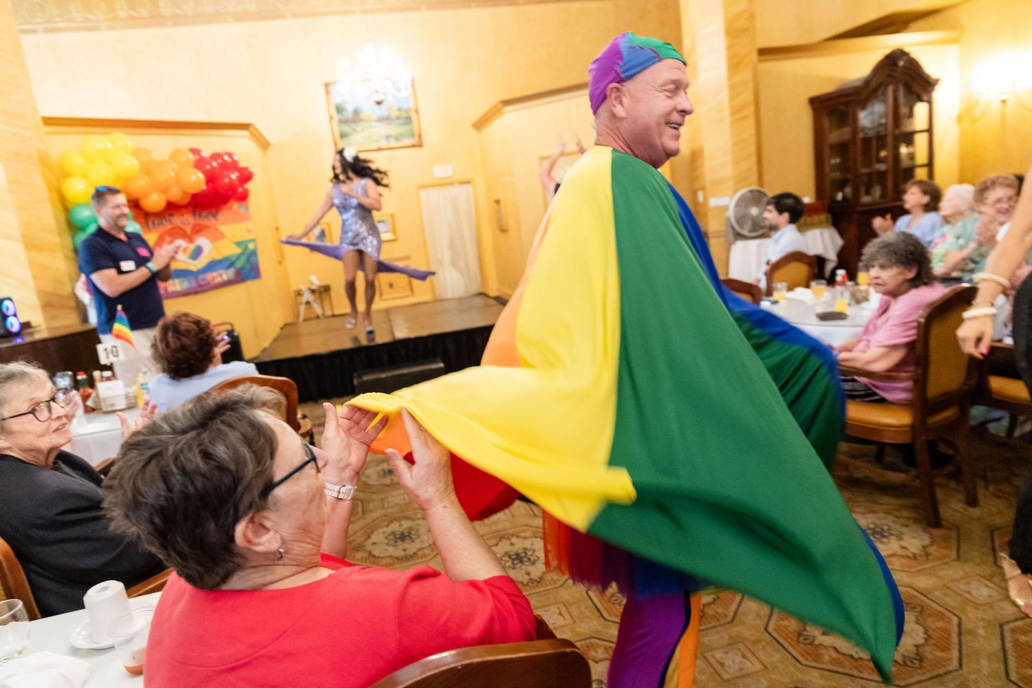 Man wearing rainbow cape interacts with seated guests at a lively Pride celebration event.