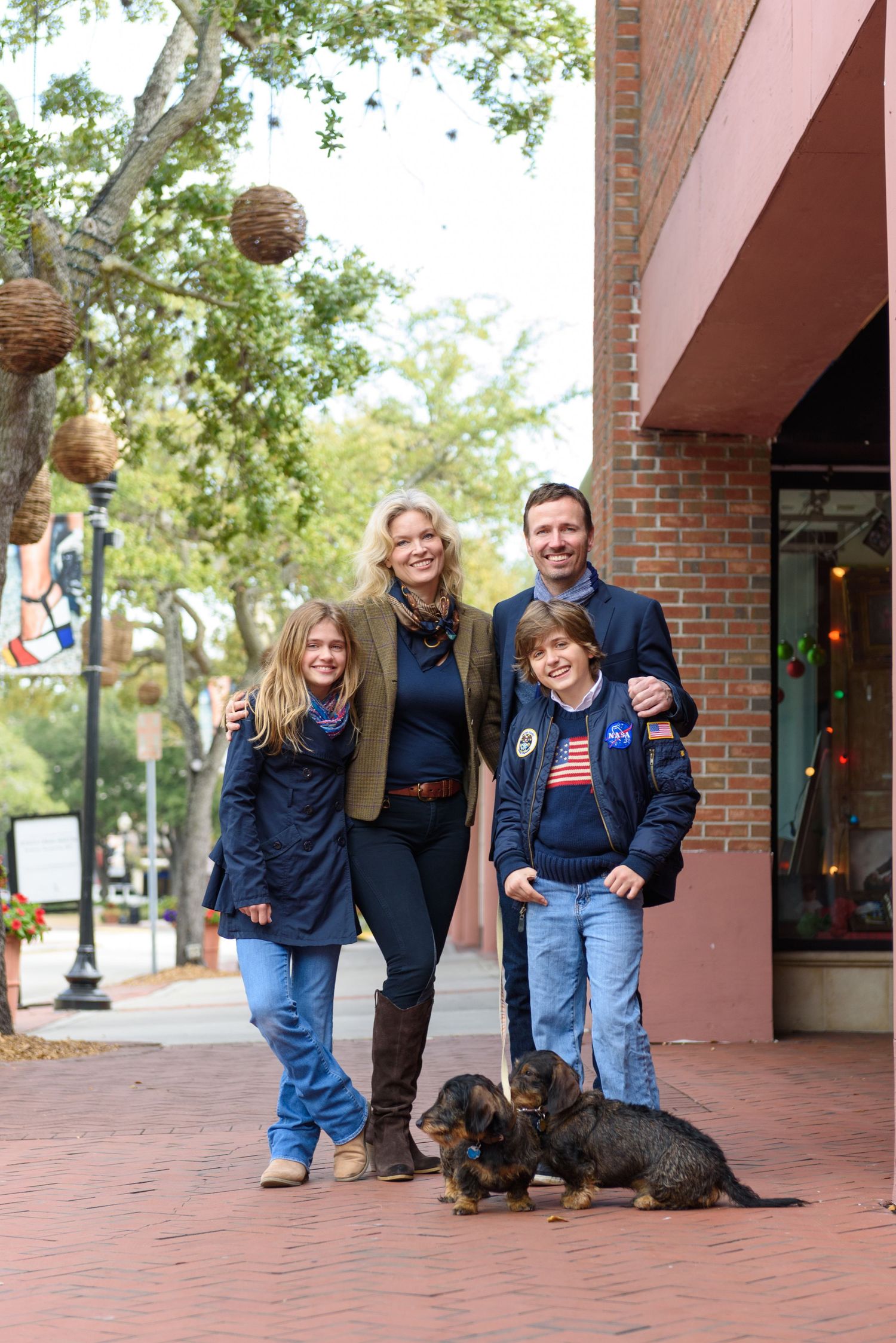 Family of four with two dachshunds posing on a brick sidewalk near shops, wearing navy blue outfits.