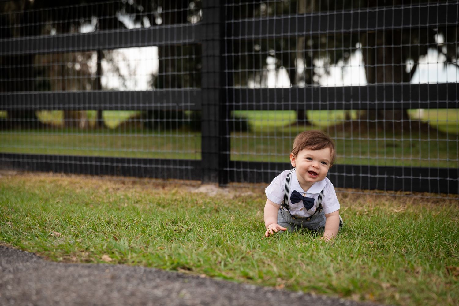 A happy baby boy crawls on green grass wearing a bow tie onesie near a black metal fence outdoors.