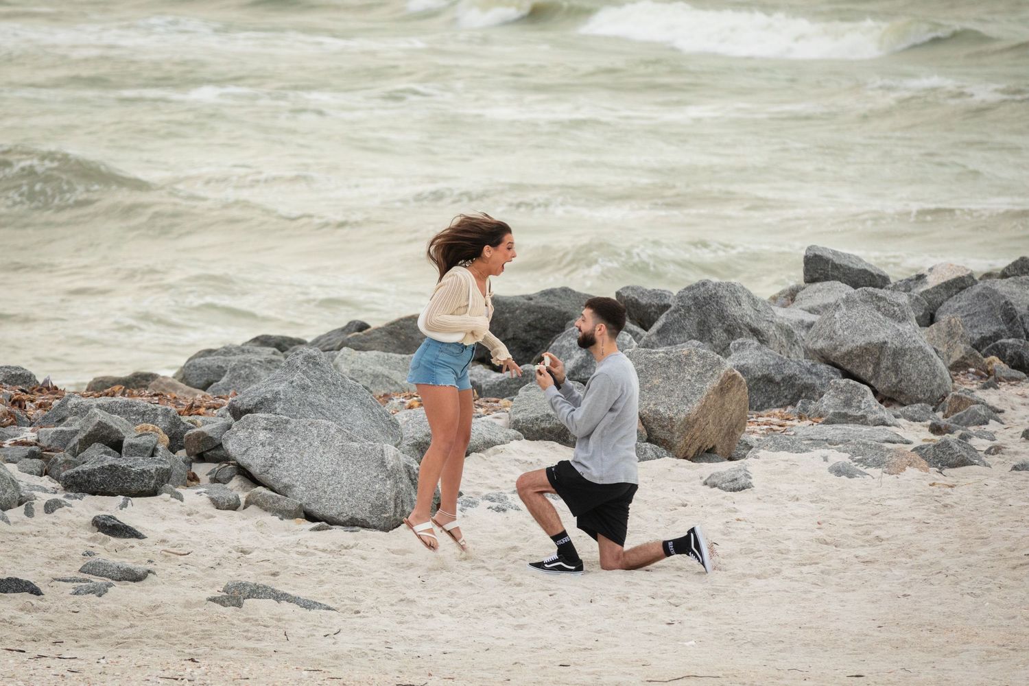 Man proposes to woman on sandy beach with rocky shoreline and ocean waves in background.