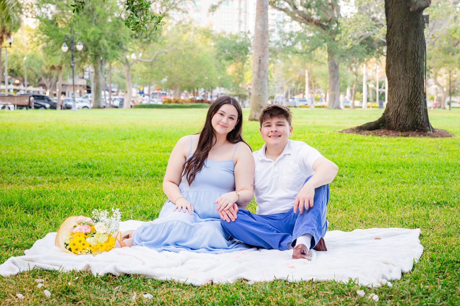Couple sitting on white blanket in park, woman in blue dress, man in white shirt, smiling at camera.