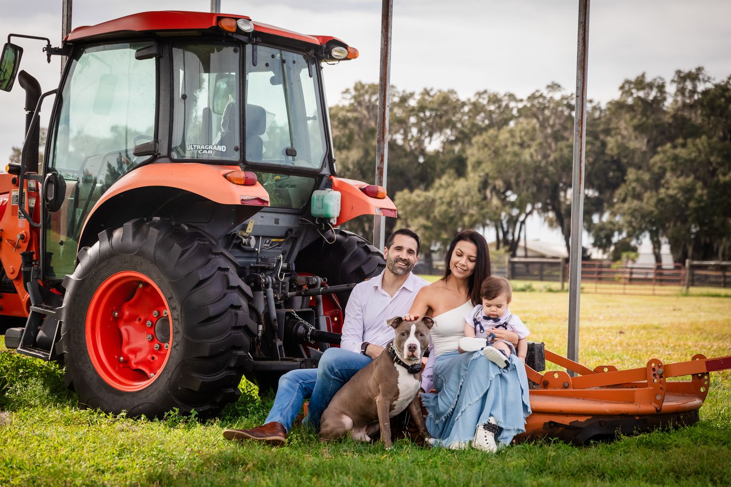 Family with dog poses beside red Kubota tractor on green farm lawn for outdoor portrait photo.