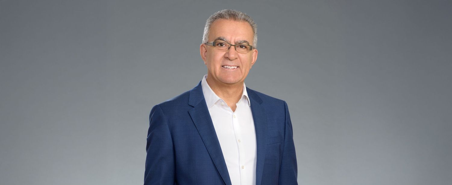 Professional man in blue blazer and white shirt poses confidently against a gray studio background.