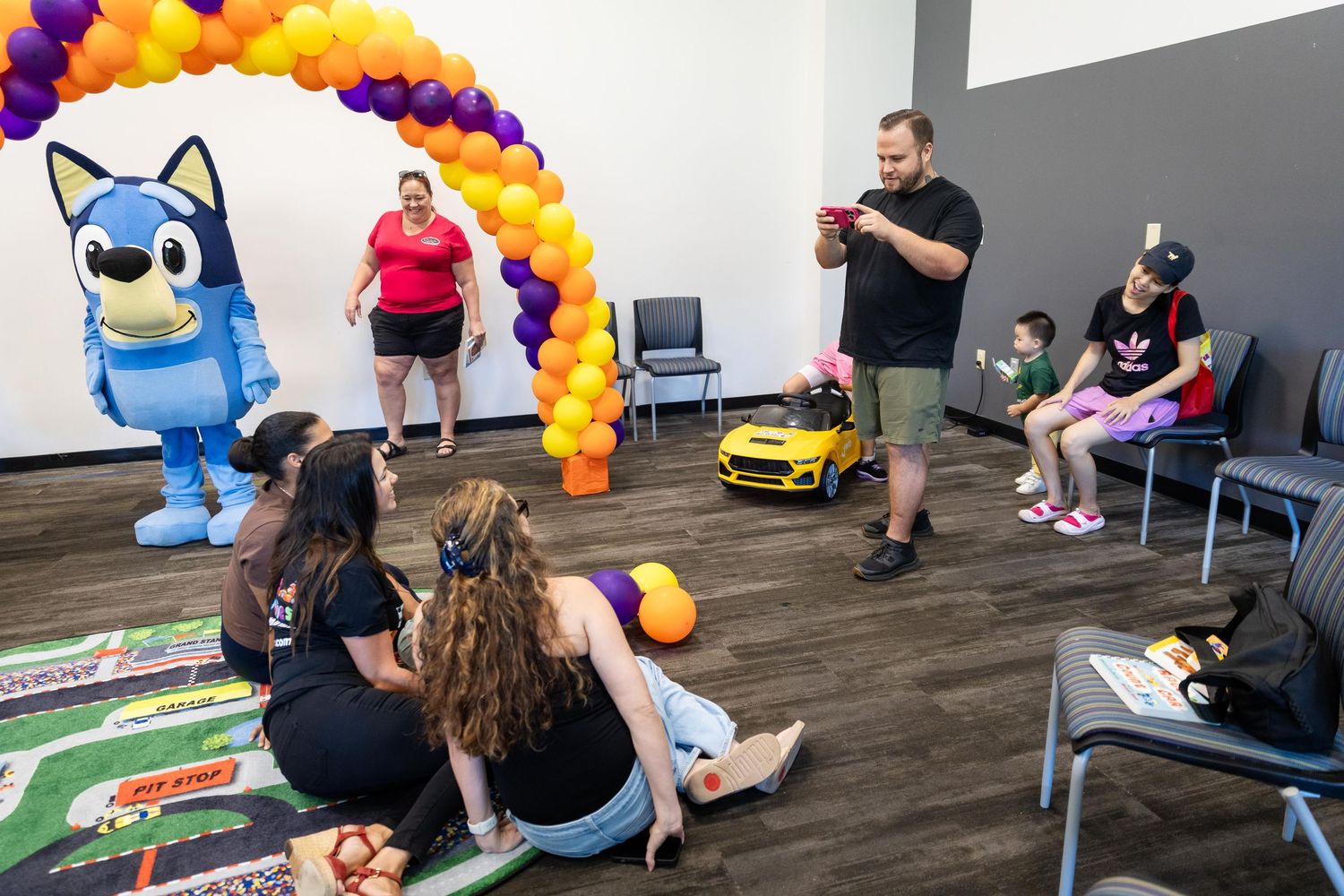 Children enjoy a Bluey themed birthday party with balloon arch, ride-on toy car, and costumed character in a bright party room.