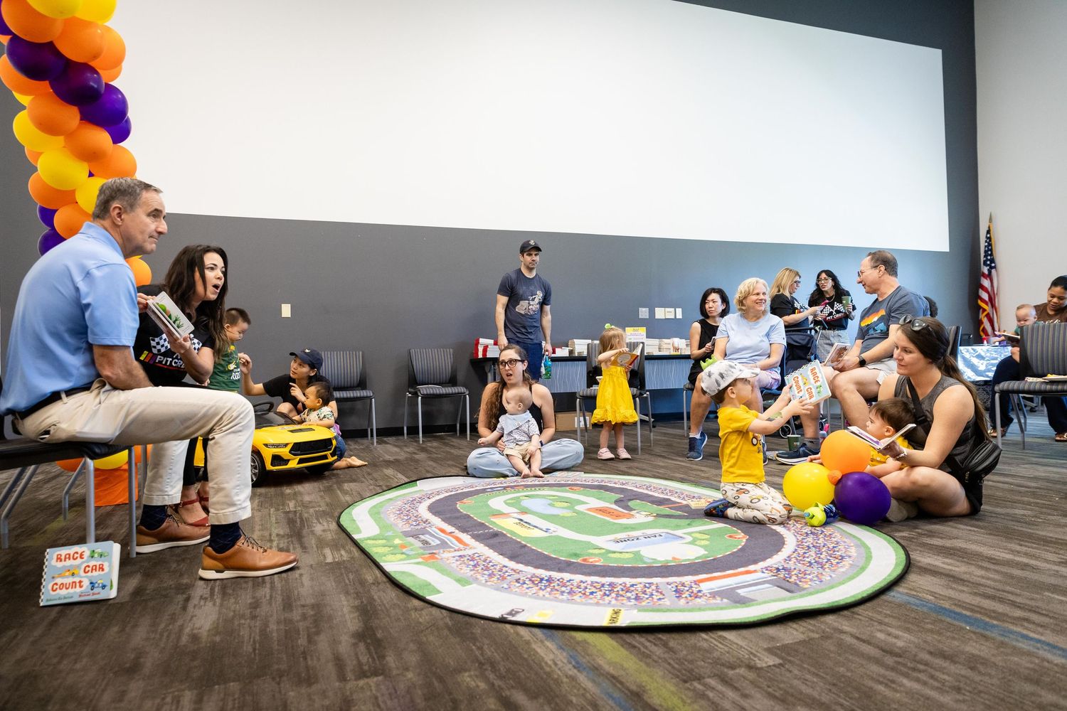 Families with young children gather in a community center for a kids event, sitting on a colorful rug with balloon decorations.