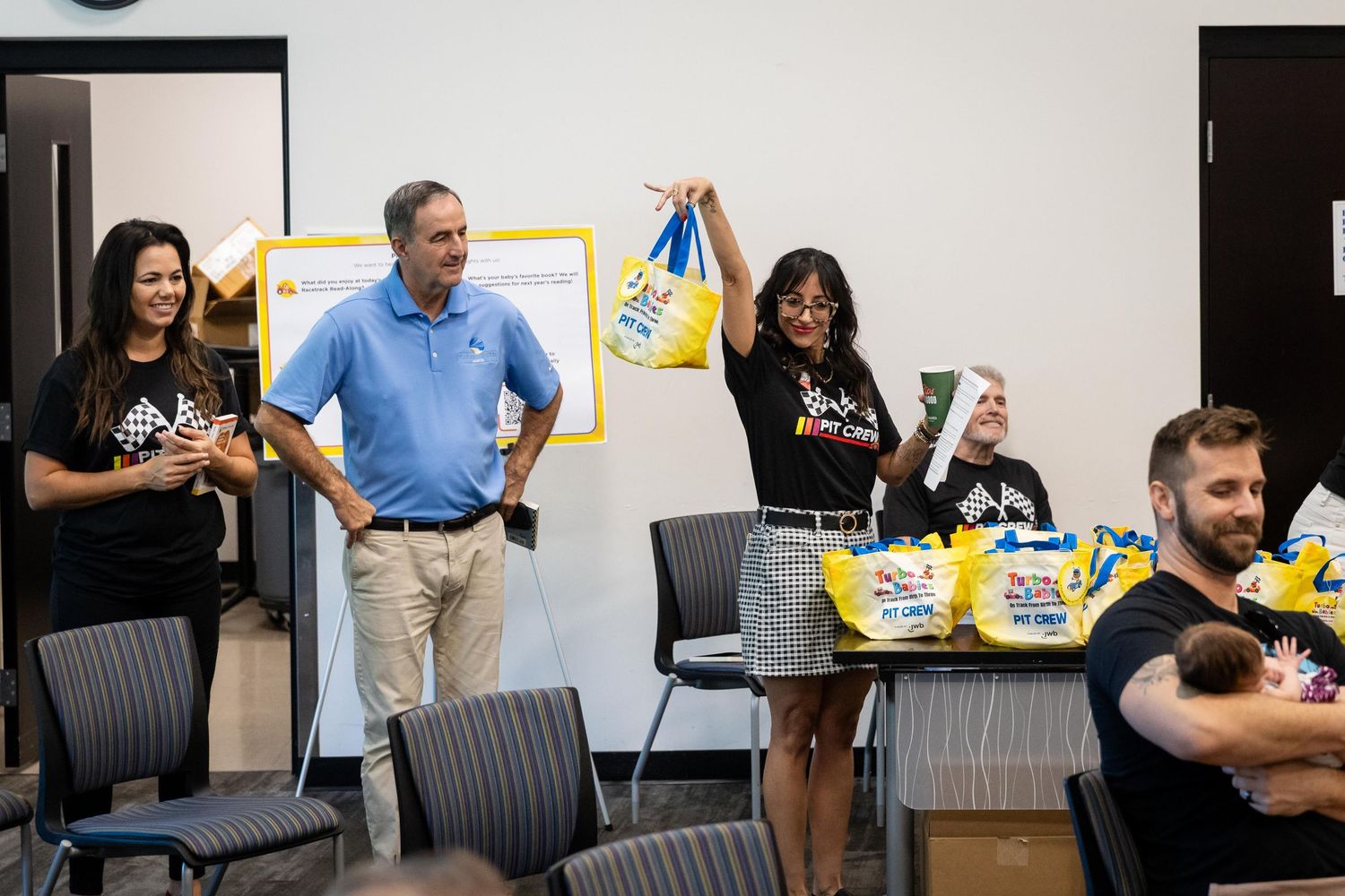 People gathered in a meeting room, woman holding colorful gift bags during a cheerful group presentation event.