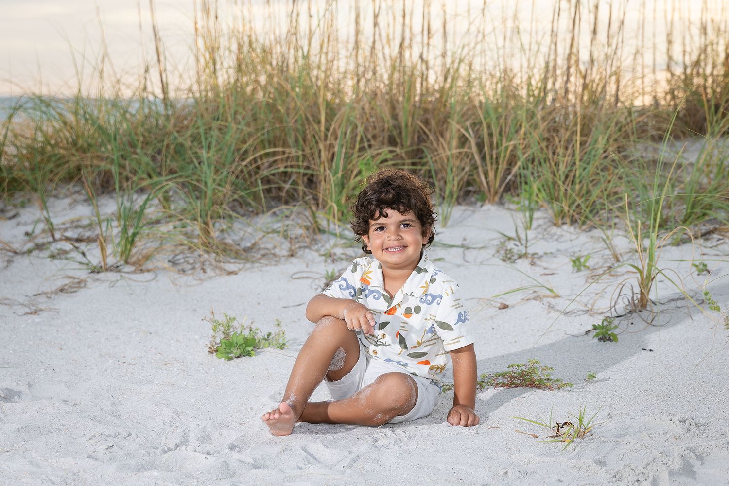 A smiling young boy sits on white sand beach dunes with sea grass in the background at sunset.