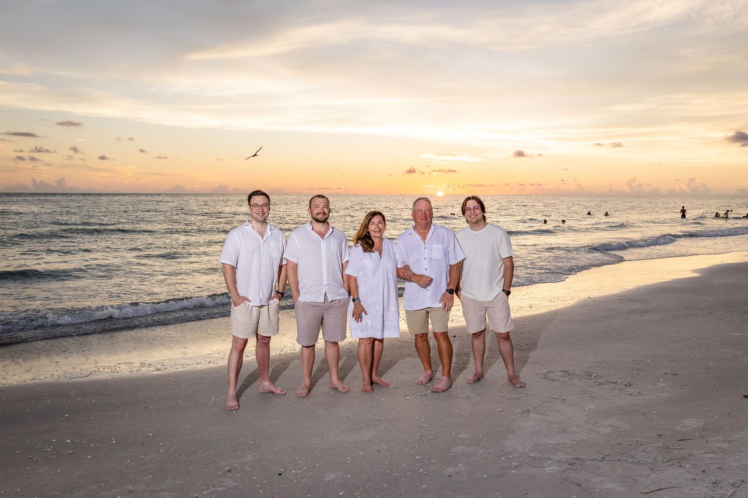 Family of five posing on a sandy beach at sunset in matching white and khaki outfits.