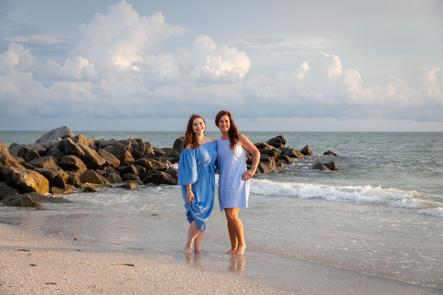Two women in blue dresses smiling on a sandy beach with rocky jetty and ocean waves behind them.