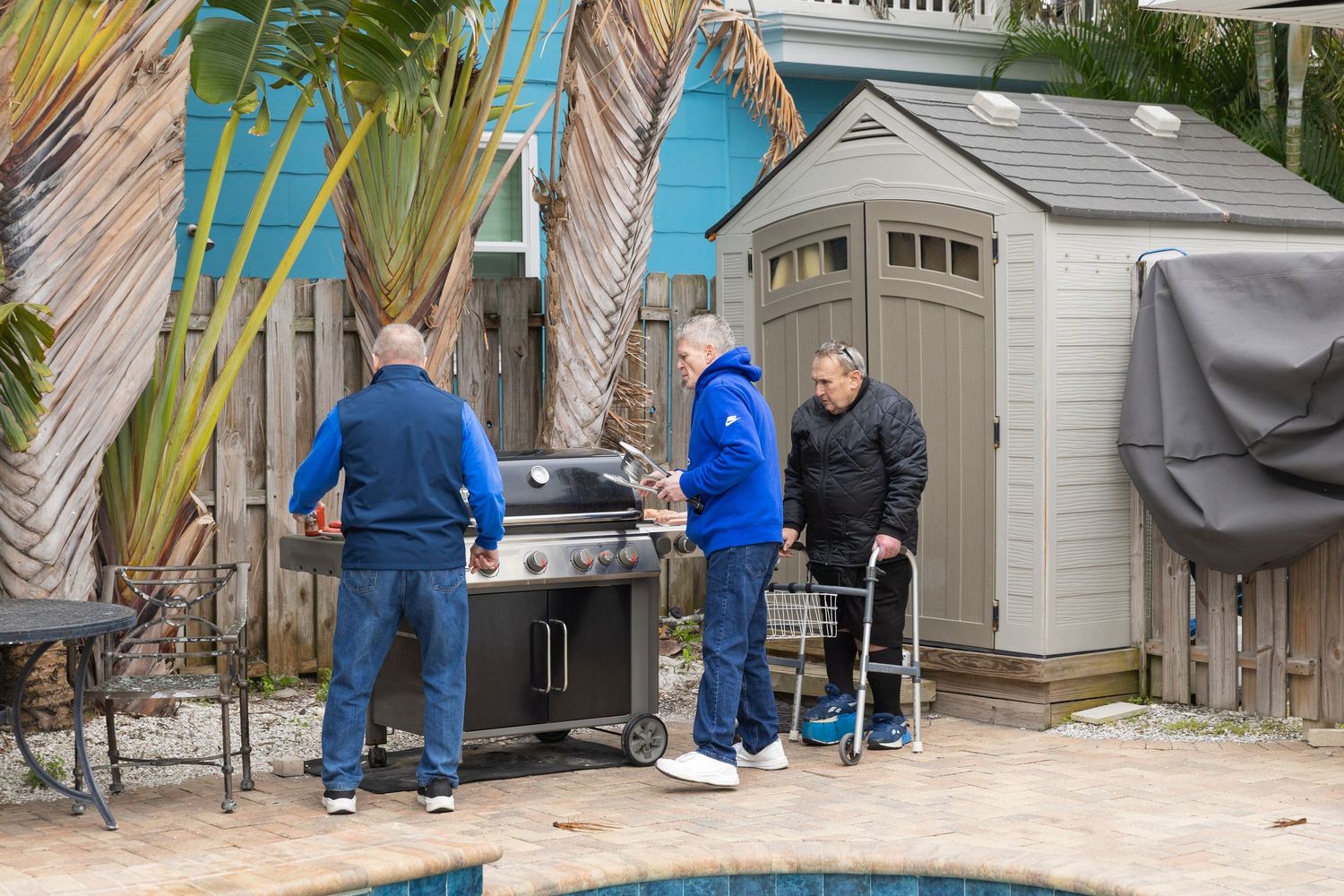 Three people grilling outdoors near a backyard pool with palm trees and a storage shed.