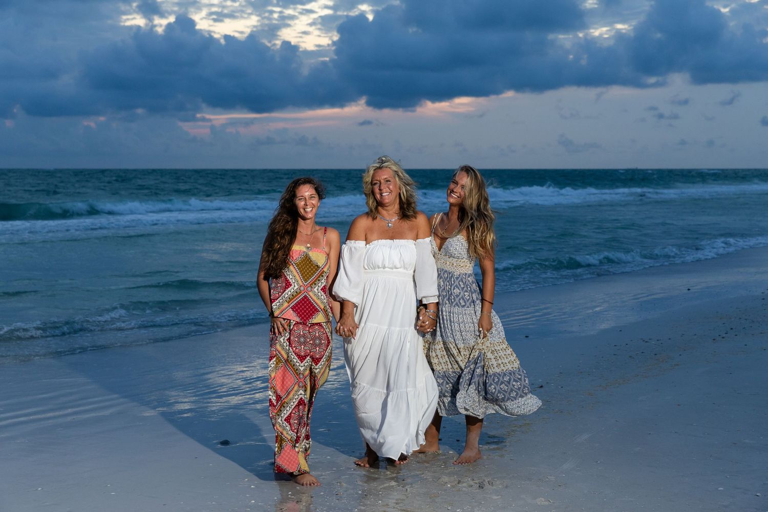 Three smiling women in boho dresses posing together on a beach at sunset with dramatic cloudy sky.
