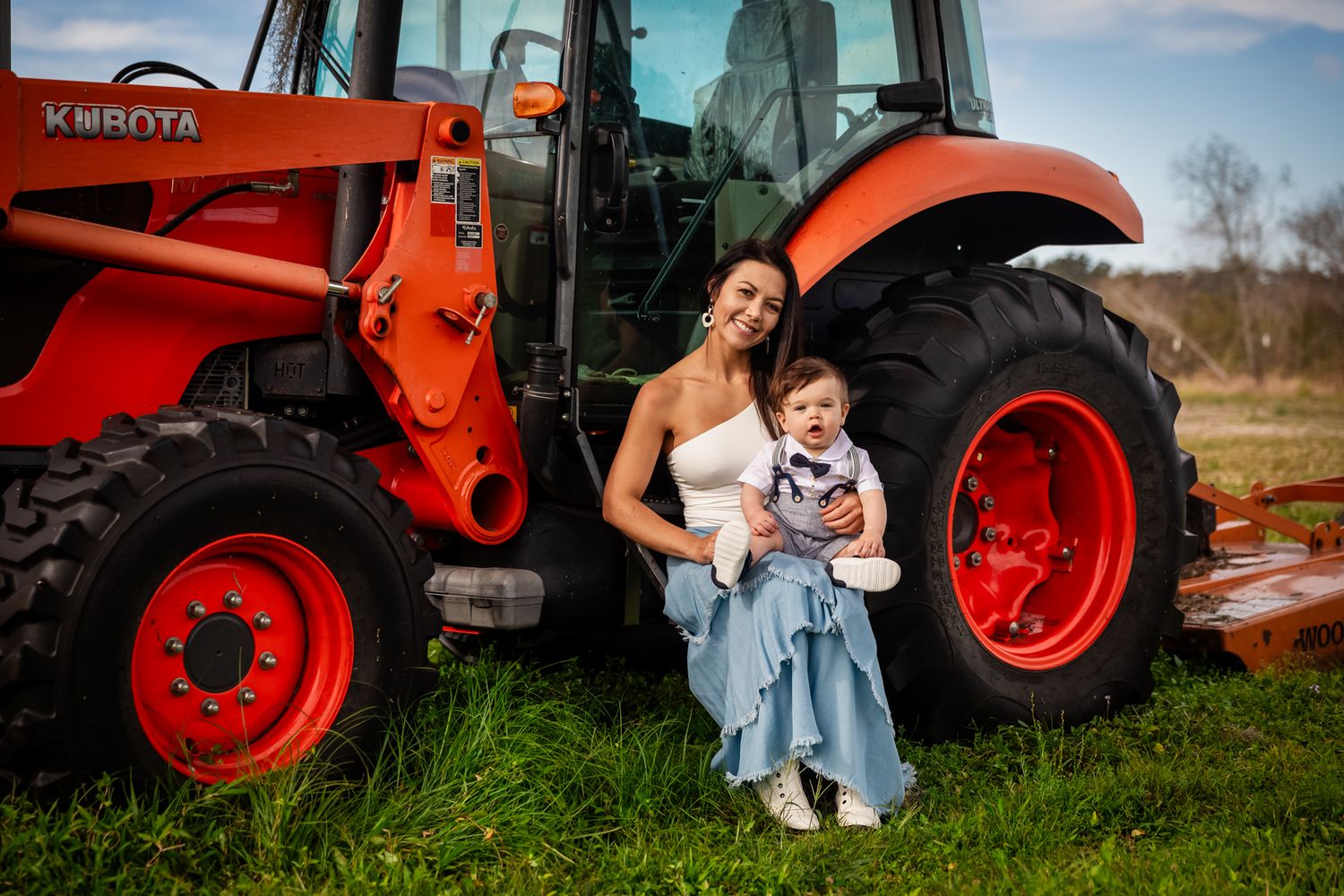 Mother and baby sitting against a red Kubota tractor on a farm, both dressed in coordinating blue and white outfits.