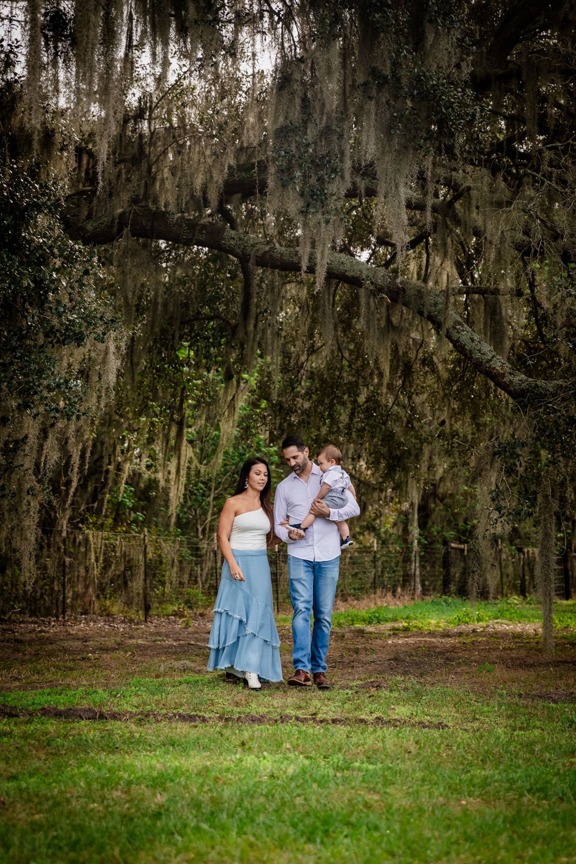 A family of three walks under Spanish moss-draped oak trees in a lush green park, with a pregnant mother in a blue skirt.