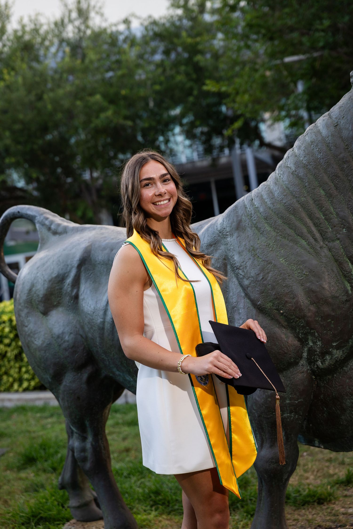 Smiling female graduate in white dress and gold sash holds cap beside bronze bull statue outdoors.
