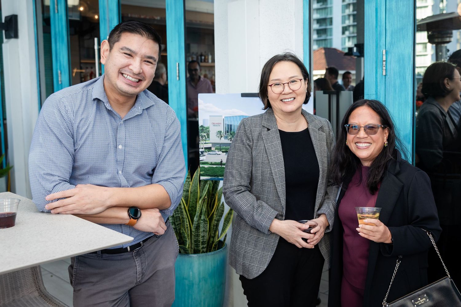 Three colleagues smile together at an outdoor networking event near teal-framed windows with a city building in background.