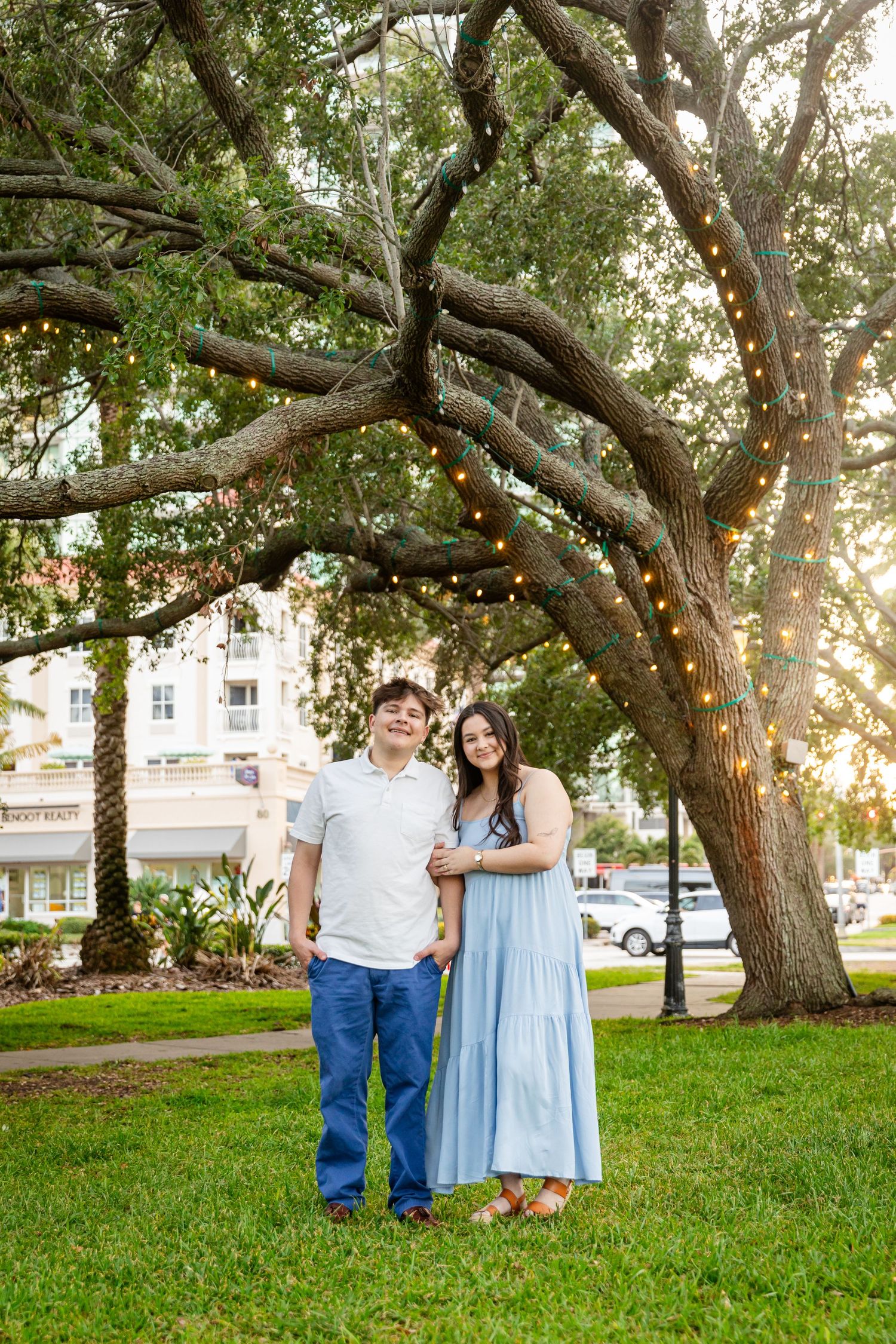 Couple standing together under large oak trees with colorful string lights in park.
