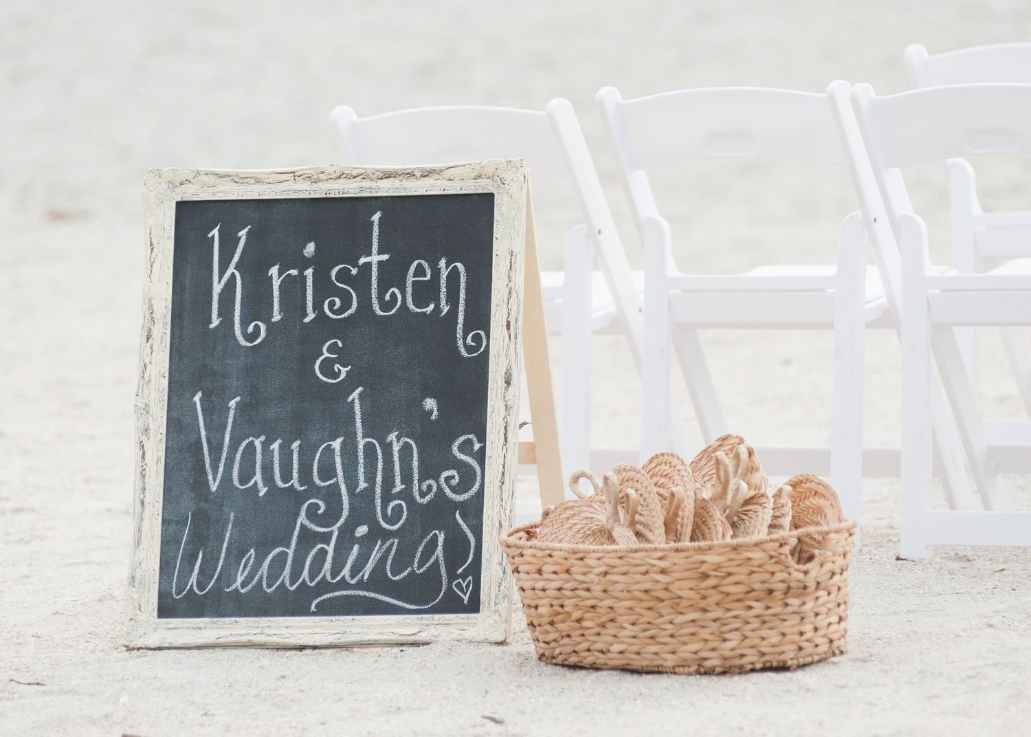Chalkboard wedding sign reading Kristen and Vaughn's Wedding beside a wicker basket and white chairs on a beach.