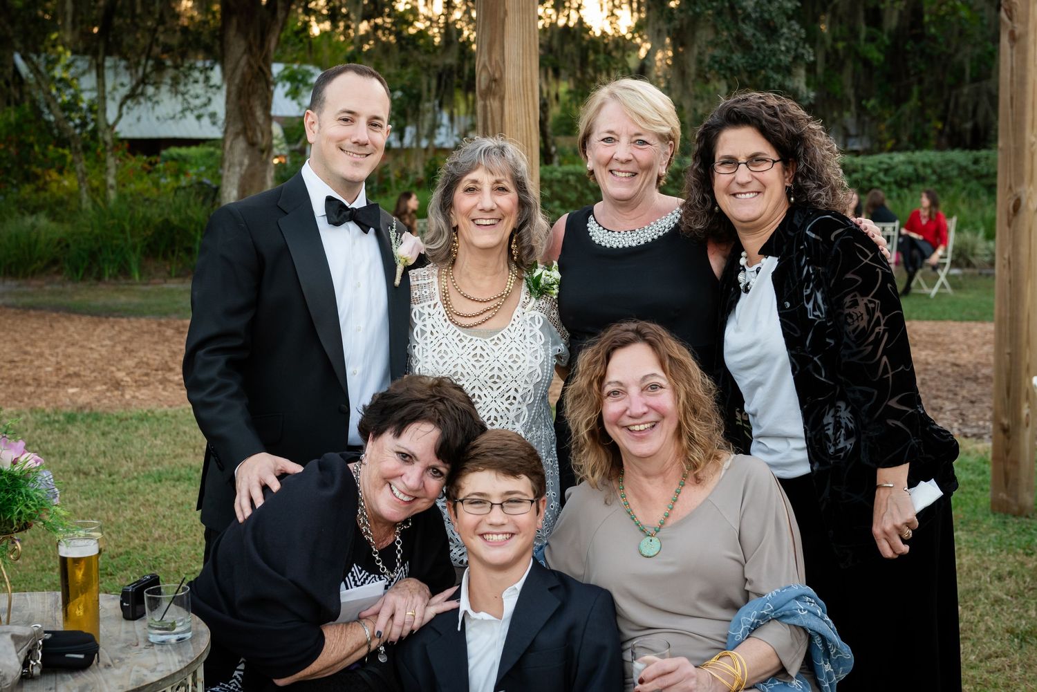 Group of seven wedding guests posing together outdoors at evening reception in formal attire.
