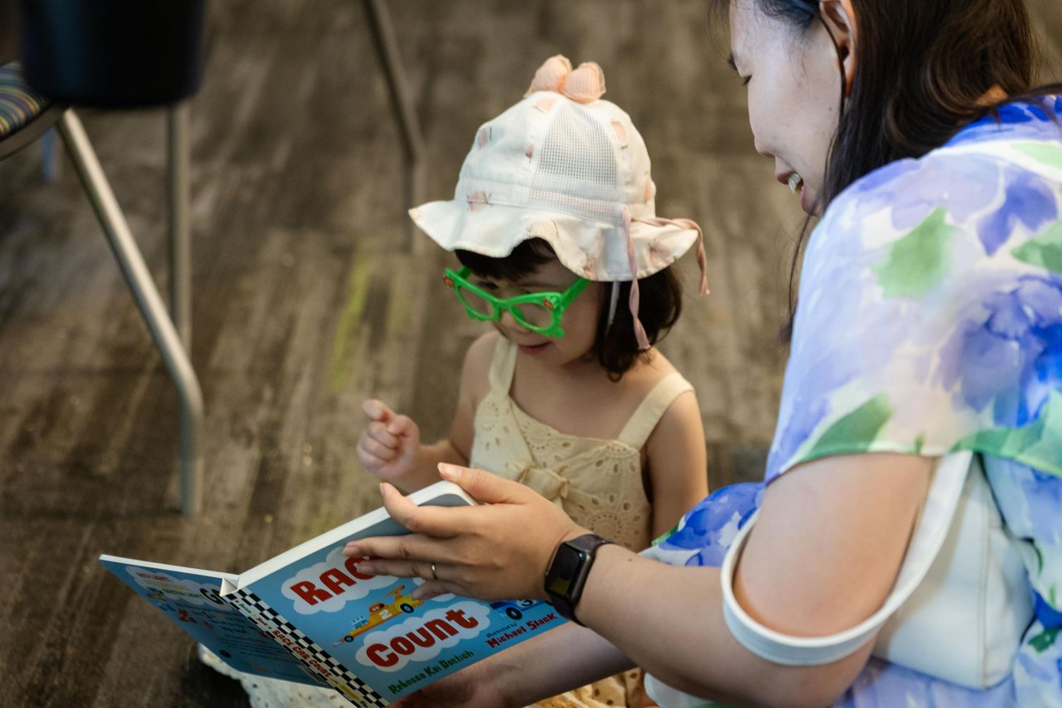 A woman and young girl in a sun hat and green glasses read a colorful children's book together on a wooden floor.