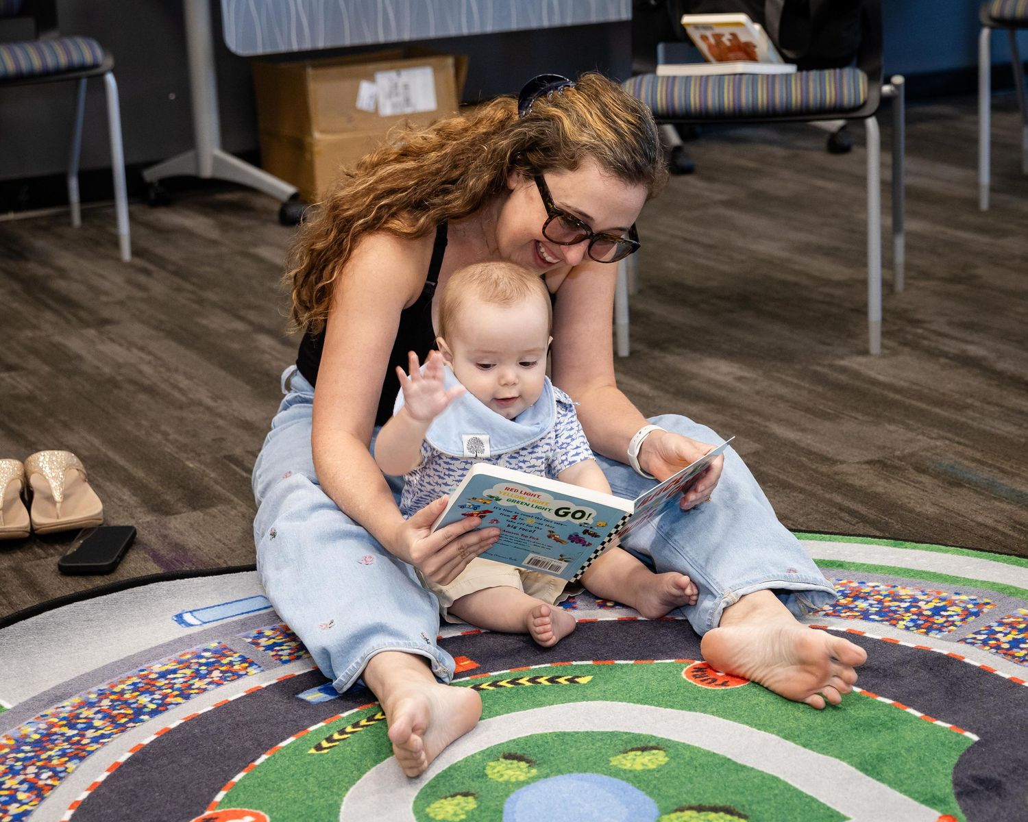 A mother sits on a colorful rug reading a picture book with her smiling baby on her lap.
