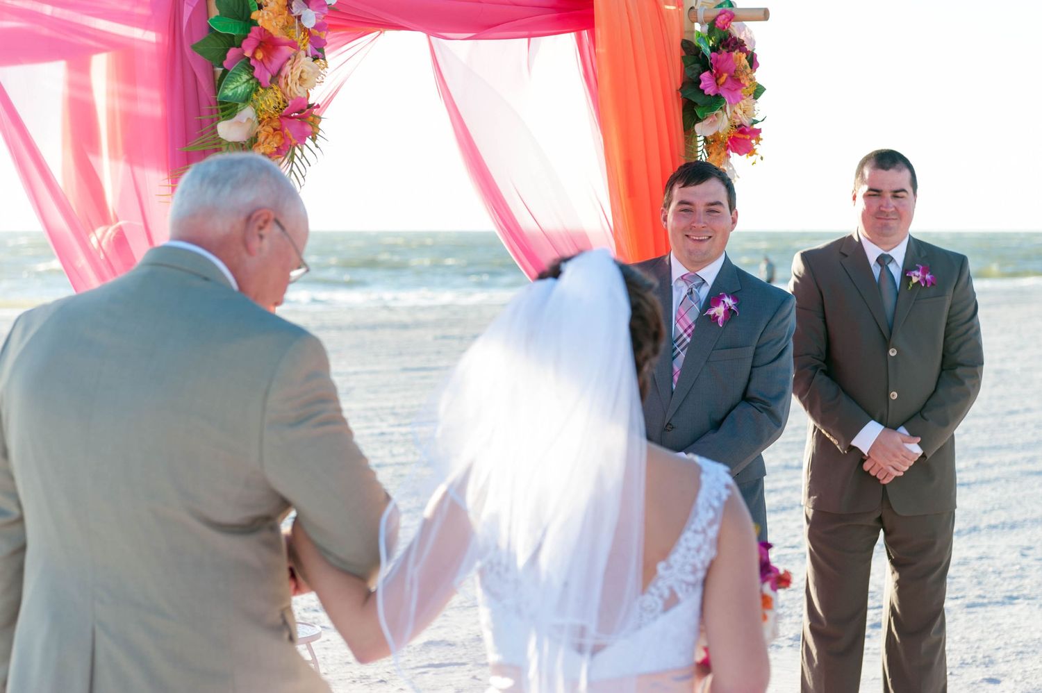 Groom smiles at bride during romantic beach wedding ceremony under colorful pink and orange floral arch.