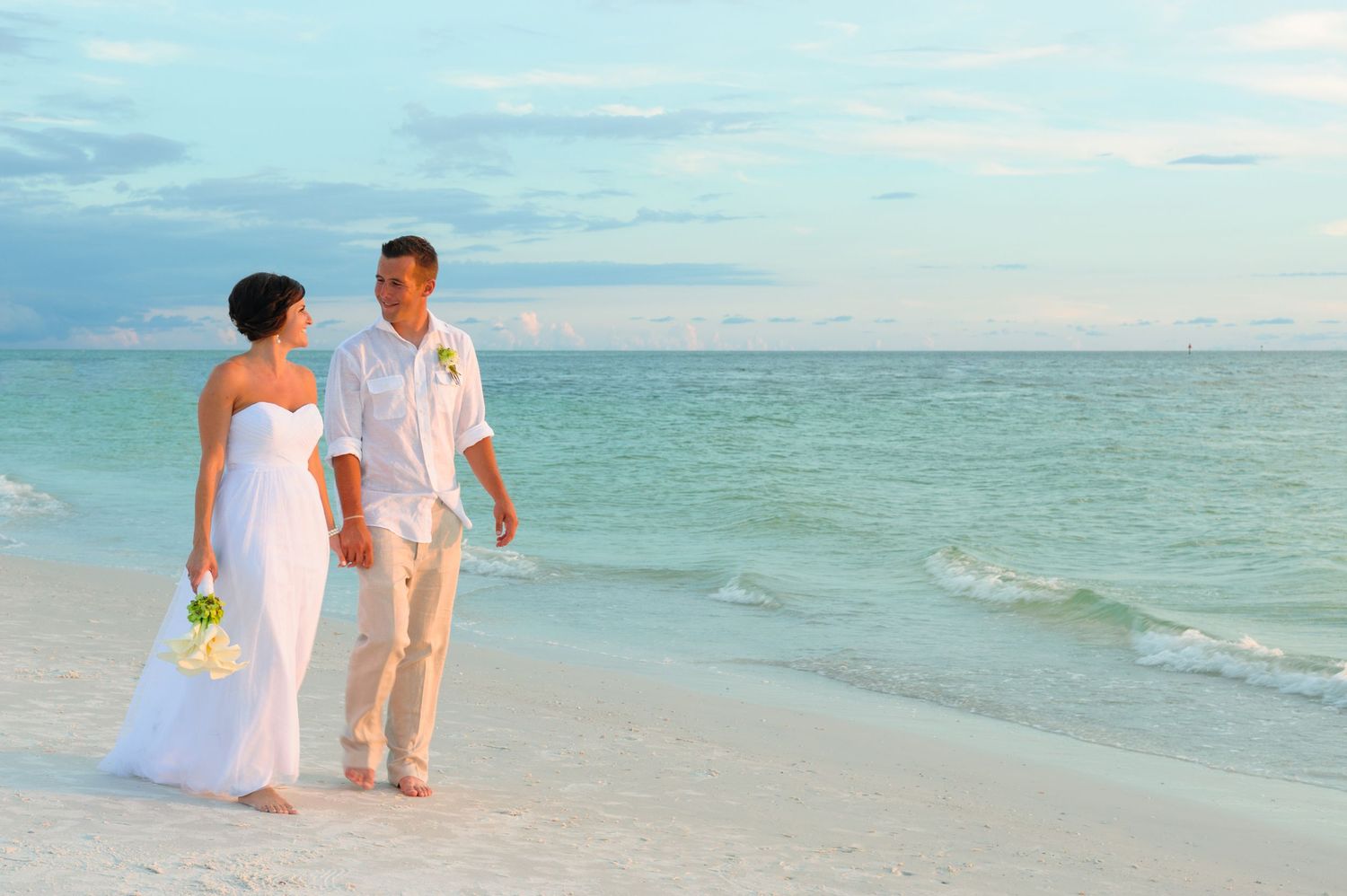 Bride and groom walking hand in hand on a white sandy beach with turquoise ocean waters.