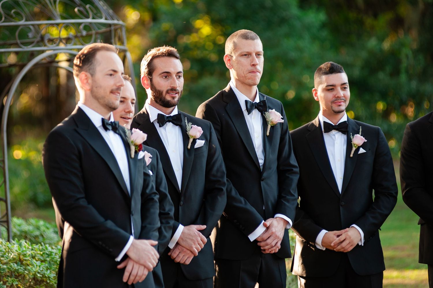 Four groomsmen in black tuxedos with boutonnieres stand attentively during outdoor garden wedding ceremony.
