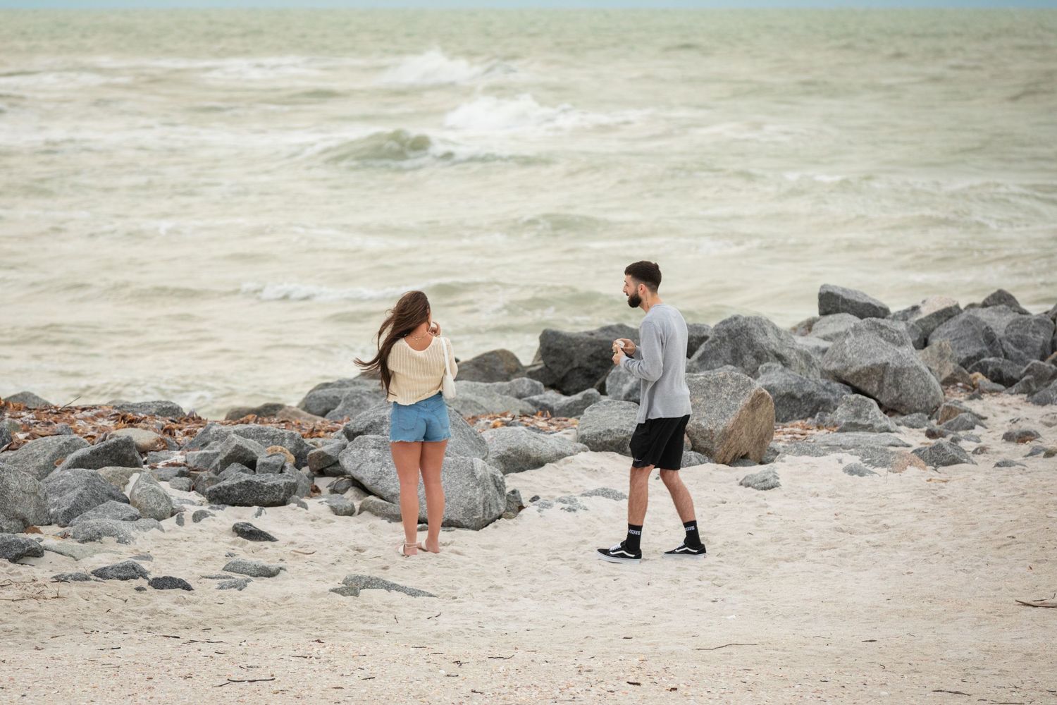 A couple walks along a rocky beach shore with waves crashing in the background on a cloudy day.
