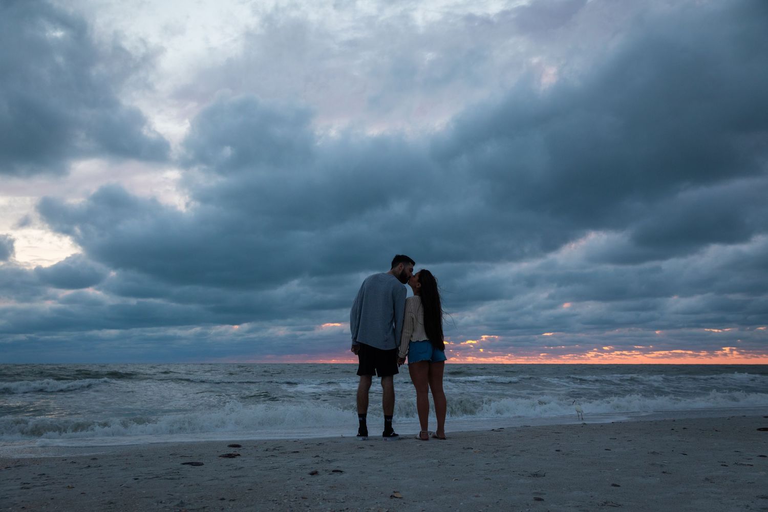 Romantic couple standing together on a beach at sunset under dramatic stormy clouds.