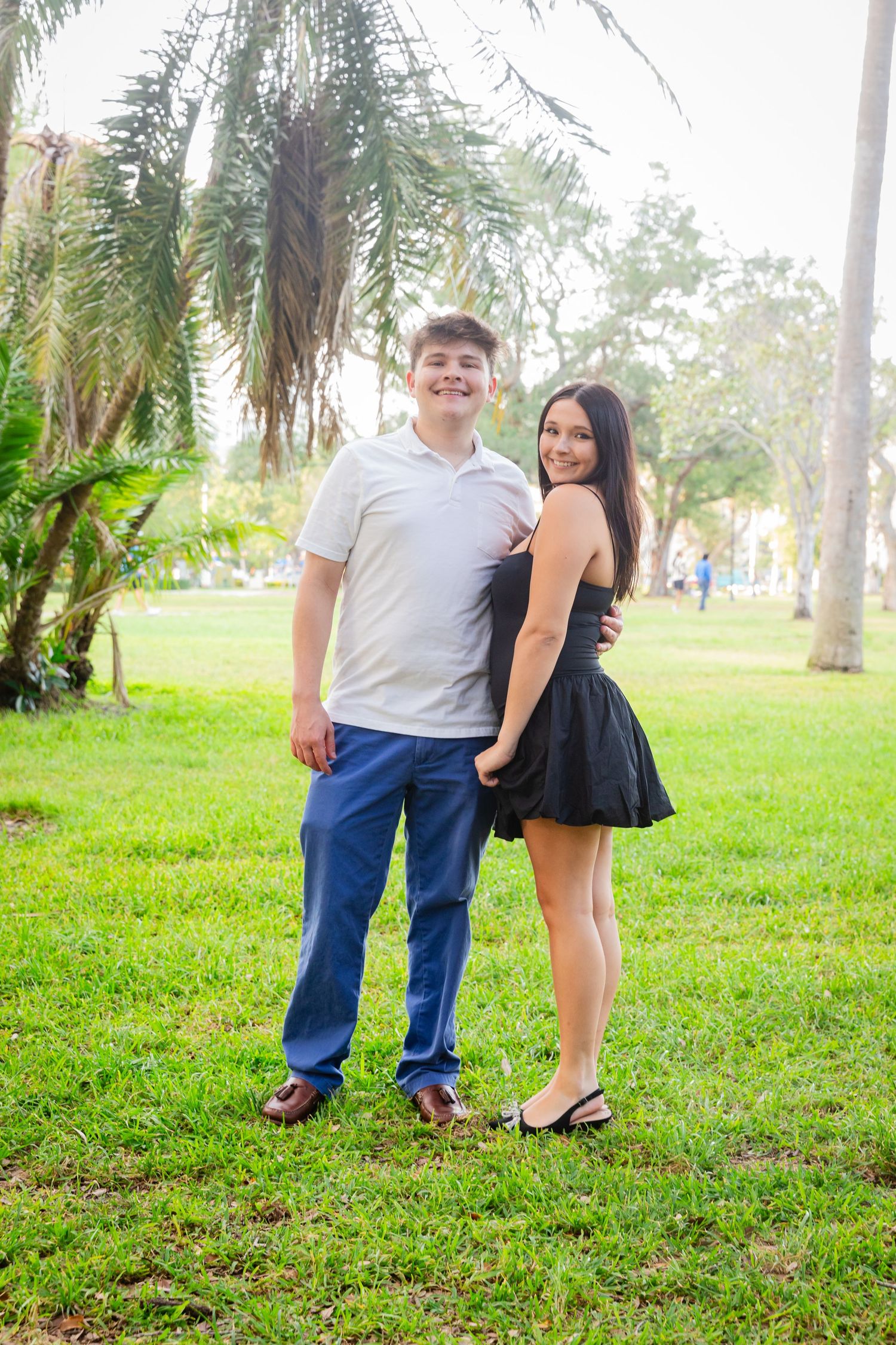 A young couple smiles together outdoors on green grass with palm trees, wearing casual attire in a tropical park setting.