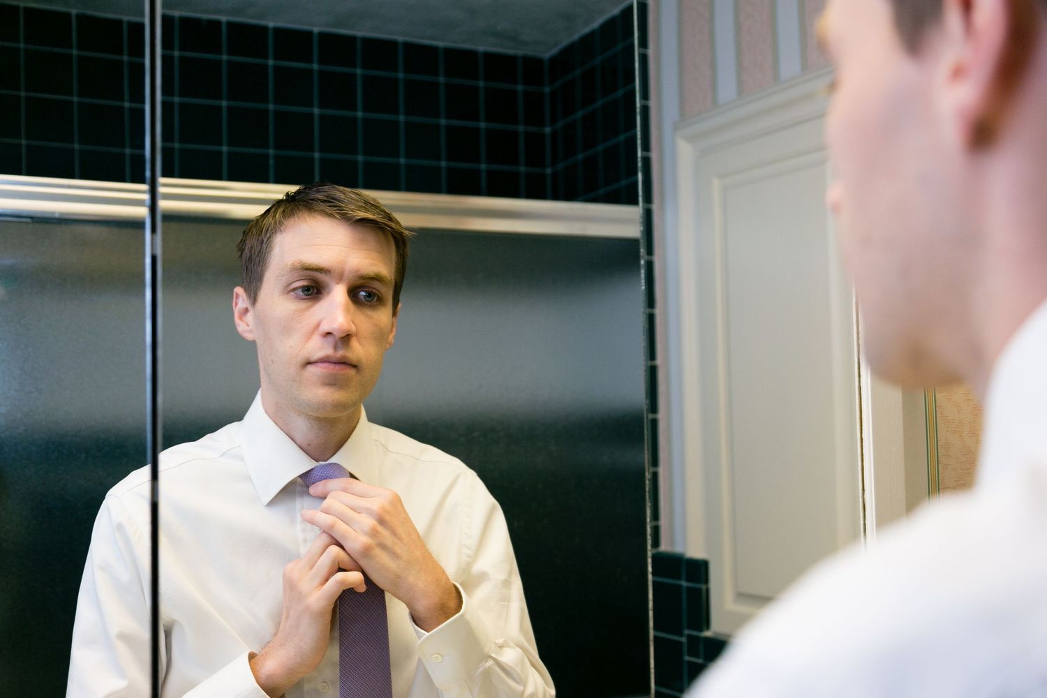Man adjusting his tie in a bathroom mirror while wearing a white dress shirt, preparing for a formal occasion.