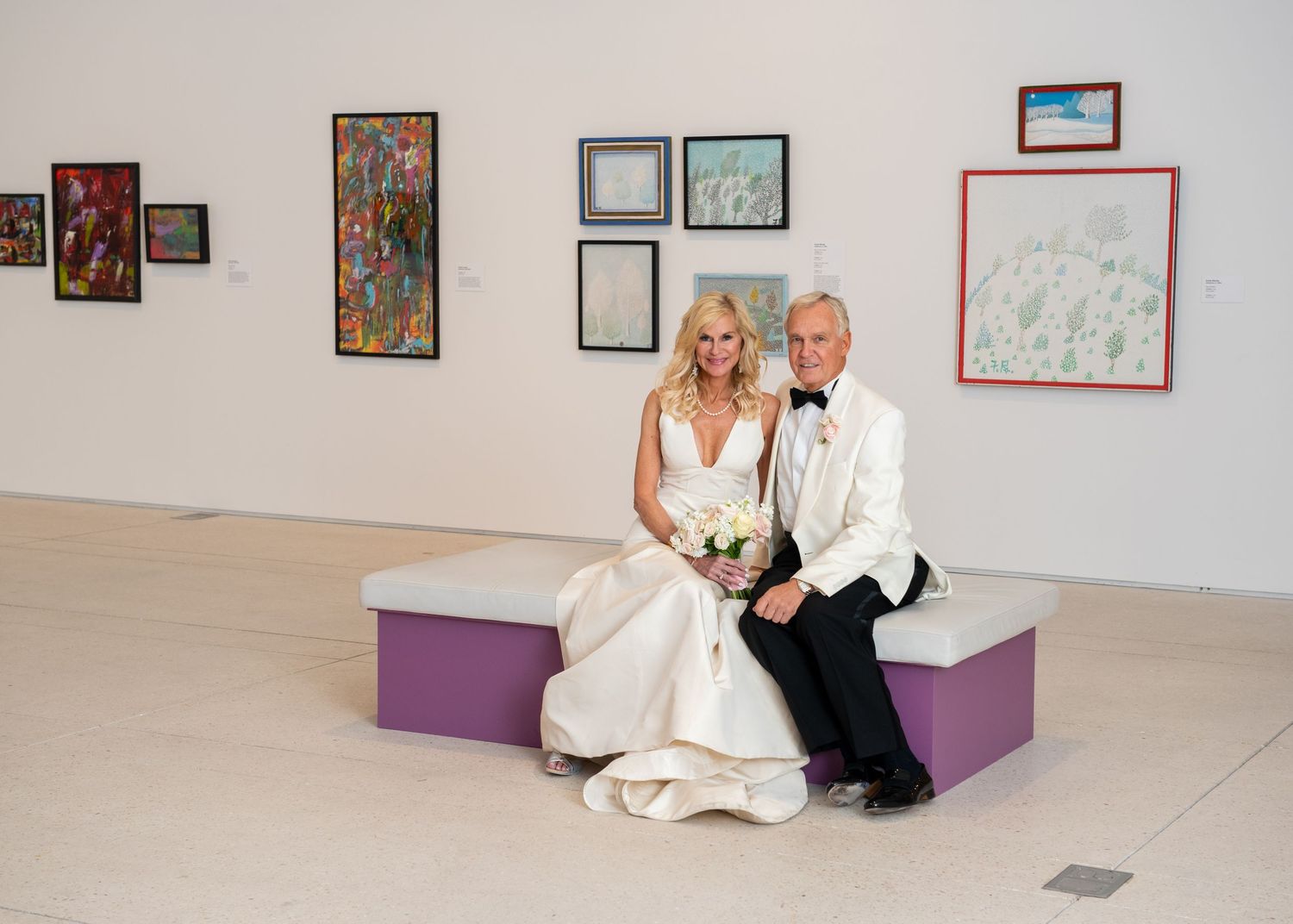 A bride and groom pose together on a bench in an elegant art gallery, surrounded by colorful paintings on white walls.